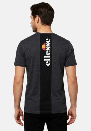 Homme portant un t-shirt gris foncé avec une bande noire verticale dans le dos affichant le logo blanc « ellesse » et un emblème orange-rouge.