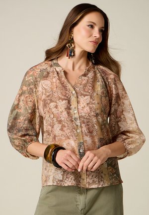 Vrouw met lang bruin haar die een blouse met bloemenpatroon en knopen draagt, grote kleurrijke oorbellen en dikke armbanden aan één pols.