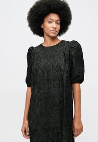 Mlada ženska s afro frizuro, oblečena v črno strukturirano obleko s kratkimi pufastimi rokavi, ki stoji pred enostavno belo ozadjem.