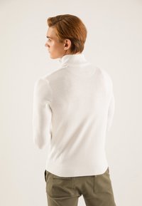 Maglione bianco a collo alto lavorato a maglia con orlo e polsini a coste, visto da dietro, abbinato a pantaloni verde oliva. Tessuto liscio, design minimalista.