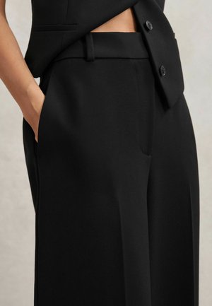 Žena nosiaca čierne na mieru šité nohavice s bočnými vreckami a zodpovedajúce čierne zapínané sako, ruka vrecku.