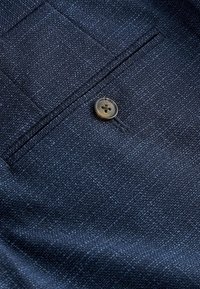 Nahaufnahme eines dunkelblauen strukturierten Stoffes mit einem einzelnen braunen Vierlochknopf neben einer horizontalen Paspeltasche.