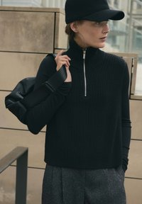 Frau trägt einen schwarzen gerippten ärmellosen Pullover mit Reißverschluss, ein schwarzes Langarmshirt, eine schwarze Kappe, graue Hosen und trägt eine schwarze Tasche über der Schulter.