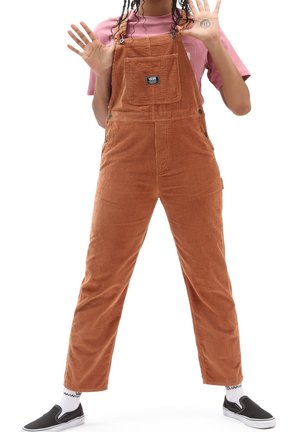 Person stående med benene spredt, iført brune kordfløjls-overalls, pink skjorte, sorte slip-on sko og viser begge hænder løftet fremad.