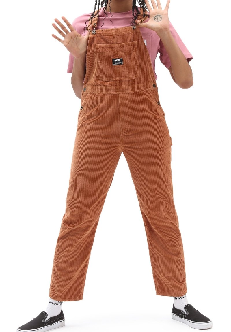Personne debout, jambes écartées, portant une salopette en velours côtelé marron, une chemise rose, des chaussures noires sans lacets, montrant les deux mains levées en avant.