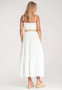 Biała sukienka maxi z warstwowym wzorem, z prążkowaną teksturą na górze i plecionym beżowym paskiem. Modelka nosi platformowe sandały z dodatkowymi detalami.