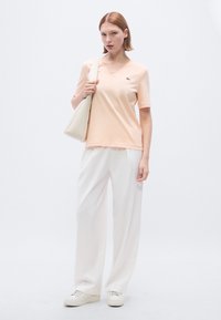 Ανοιχτό ροδακινί T-shirt με V λαιμόκοψη από απαλό ύφασμα, συνδυασμένο με φαρδιά λευκά παντελόνια και λευκά αθλητικά παπούτσια. Διαθέτει μικρό λογότυπο στο στήθος.