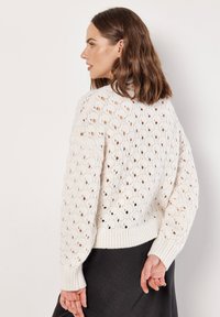 Cremefarbener Kabelstrickpullover mit offenem Muster, gerippten Bündchen und Saum, entspannter Schnitt und weicher Textur.