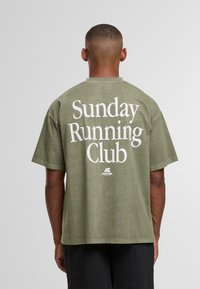 Muž s krátkými vlasy, oblečený v olivově zeleném tričku s nápisem "Sunday Running Club" vytištěným bílou barvou na zadní straně, stojící na jednobarevném pozadí.