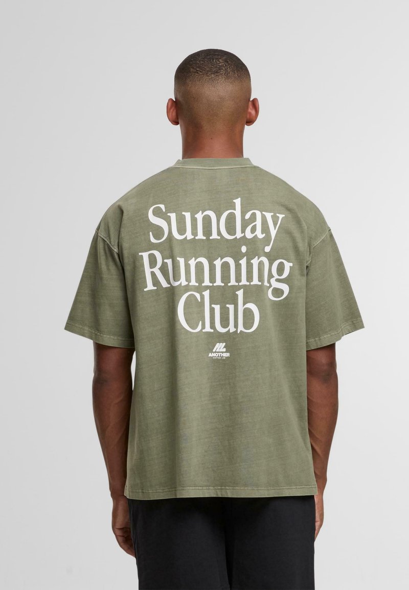 Muž s krátkými vlasy, oblečený v olivově zeleném tričku s nápisem "Sunday Running Club" vytištěným bílou barvou na zadní straně, stojící na jednobarevném pozadí.