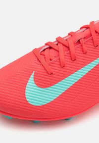 Scarpa da calcio sintetica rossa con superficie testurizzata, caratterizzata da un grande logo Nike aqua e da lacci e tacchetti vibranti a contrasto.