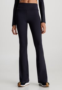 Leggings neri a zampa d'elefante realizzati in materiale elastico con un alto punto vita. Texture liscia e vestibilità su misura, abbinati a scarpe da ginnastica scure.