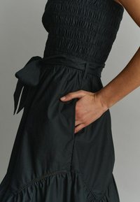 Musta olkaimeton kesämekko, jossa on smokkattu yläosa, solmittava vyö ja tasku; helma on röyhelöity ja koristettu ompeleilla.