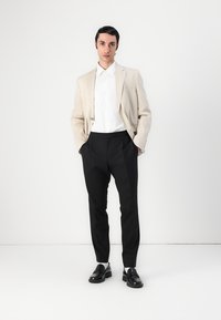 Blazer beige chiaro sopra una camicia bianca, abbinato a pantaloni neri e mocassini neri. Tessuti morbidi, vestibilità su misura, semplice dettaglio con bottone sul blazer.