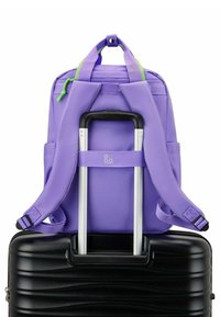 Zaino viola realizzato in tessuto resistente, dotato di doppie spalline, una maniglia superiore e zip di accento verdi. Appoggiato su una valigia nera con ruote.