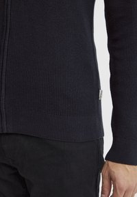 Marineblå strikket cardigan med ribbestrikket kant og mansjetter; har et sidemerke. Teksturen er jevn, med en tettsittende design.