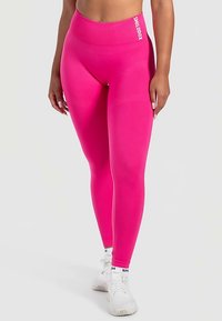 Leggings rosa aderenti con vita alta e texture a coste. Presentano un logo bianco sulla cintura. Abbinati a scarpe sportive bianche.