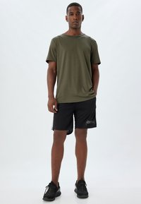 Olijfgroene shirt met korte mouwen, gecombineerd met zwarte sportshorts met witte accenten en een afdrukdetail op het been. Zwarte sneakers.