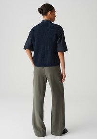 Top senza maniche blu navy a trama con collo alto indossato sopra una camicia scura a maniche corte, abbinato a pantaloni larghi verde oliva e calzature nere.