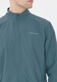 Teal lättviktsoverall, hög krage, hel dragkedja fram, "ENDURANCE"-logotyp i vitt, slät textur, rena linjer utan synliga mönster.