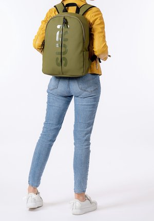 Person trägt eine senffarbene Jacke, hellblaue Jeans, weiße Turnschuhe und einen olivgrünen Rucksack mit großem vertikalem Text.