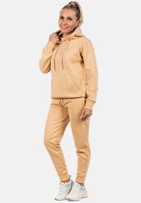 Set di felpa e pantaloni jogger color cammello, realizzato in tessuto morbido, con tasca frontale, cappuccio con cordini e polsini elasticizzati. Abbinato a sneaker bianche.