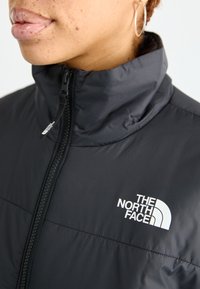 Giacca nera isolata con colletto alto, chiusura con zip e logo bianco. Materiale liscio e leggero con texture trapuntata.