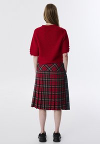 Roter kurzärmeliger Strickpullover mit dekorativen Knöpfen, kombiniert mit einem plissierten Karorock in Rot-, Navy- und Beigetönen; schwarze Sneakers.