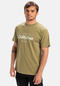 Oliven grøn bomulds t-shirt med rund med halsudskæring, prydet med hvidt trykt logo "Quiksilver" og tekst. Korte ærmer og afslappet pasform.