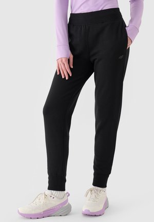 Persona que lleva pantalones jogger negros, camiseta de manga larga morada y zapatillas blancas con detalles morados, de pie con una mano en el bolsillo.