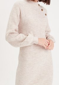 Robe en tricot beige clair, avec un col montant, une texture côtelée et trois boutons décoratifs dorés sur l\'épaule.
