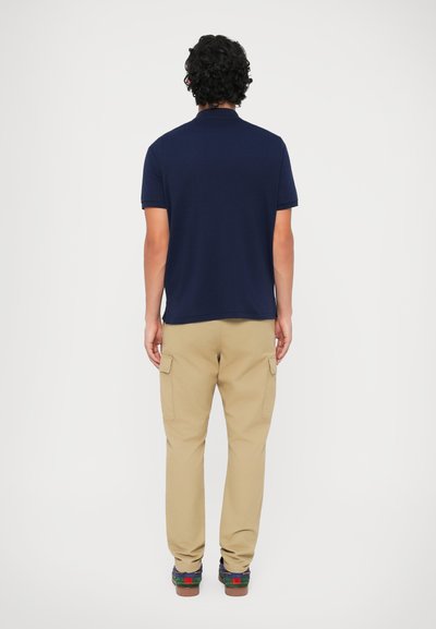 Mornarska polo majica s kratkimi rokavi, bež cargo hlače s stranskimi žepi in večbarvni čevlji. Opazna sta gladka tekstura in sproščen kroj.