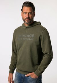 Khaki groene hoodie van een zachte stof, met een grote grijze opdruk die "HERITAGE DRY GOODS" op de borst zegt en een koord in de capuchon.