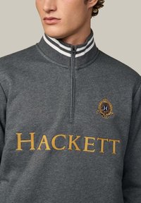 Grijze sweatshirt met hoge kraag, halve rits, witte gestreepte accenten en gouden "HACKETT" logo. Heeft een embleem met geborduurde details.
