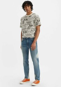 Camiseta gráfica de manga corta con estampado de periódico en color crema, combinada con jeans desgastados en azul claro y zapatillas naranjas, con un escote redondo.