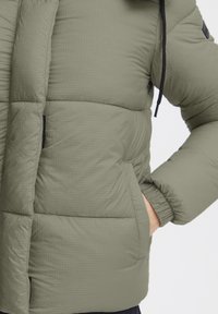 Grønn puffjakke med rutete tekstur, som har en glidelåslomme, elastiske mansjetter og hette-design.