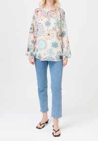 Blouse légère et transparente avec des motifs mandala multicolores, col rond avec un lien, manches longues ; associée à un jean bleu clair et des sandales noires.