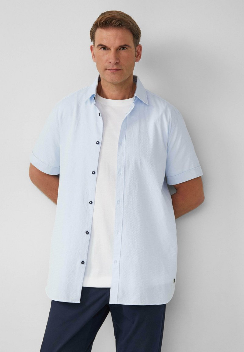 Camicia azzurra chiara a maniche corte con bottoni neri, in tessuto testurizzato e dalla vestibilità comoda, indossata sopra una maglietta bianca.