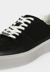 Basket en cuir noir avec une semelle blanche épaisse et des lacets blancs passés à travers une étiquette en métal sur la languette, montrant les détails latéraux et supérieurs.
