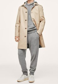 Beige regenjas met bruine knopen, gedragen over een grijze hoodie en bijpassende joggingbroek. Compleet met witte sneakers op een neutrale achtergrond.