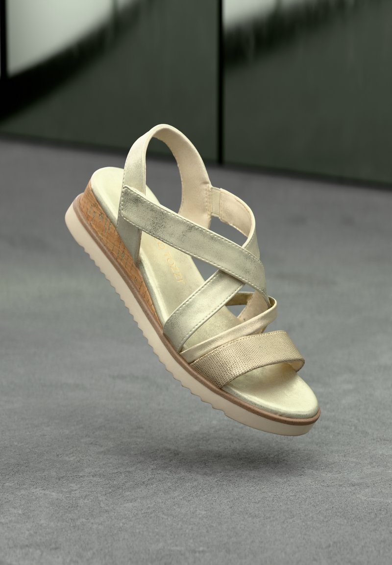 Sandale beige et dorée avec des lanières croisées, des accents texturés, une semelle intermédiaire de type liège et une semelle extérieure en caoutchouc, flottant au-dessus d'une surface grise.