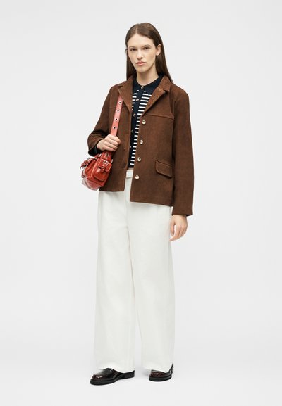 Γυναίκα με ίσια καστανά μαλλιά που φοράει καφέ μπουφάν πάνω από ριγέ πουκάμισο, λευκό παντελόνι με φαρδύ πόδι, μαύρα παπούτσια και κρατάει κόκκινη τσάντα στον ώμο.