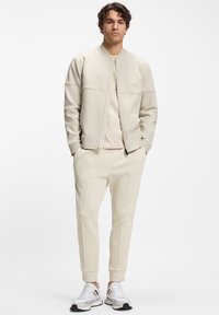 Lichtbeige sportief pak gemaakt van zachte stof, bestaande uit een bomberjack met ritssluiting en taps toelopende broek met naaddetails. Gecombineerd met witte sneakers.