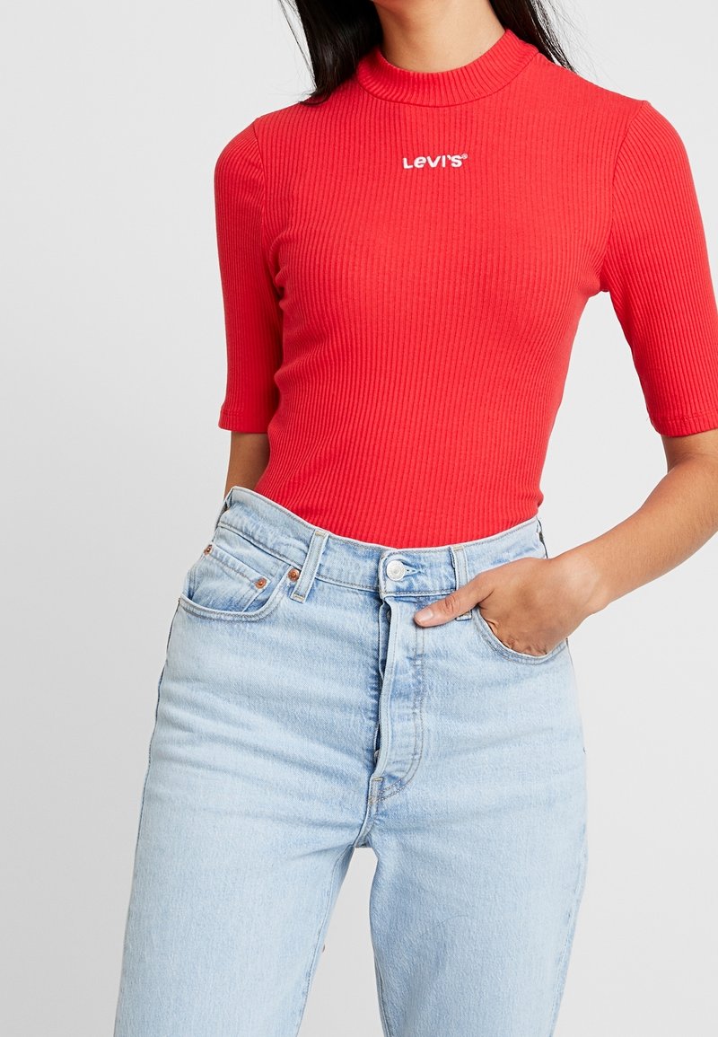 Červený žebrovaný krátký rukáv bodysuit s vysokým výstřihem; má bílé logo "LEVI'S". V kombinaci se světle modrými džínami s vysokým pasem.