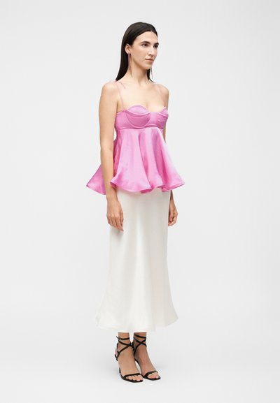 Γυναίκα που φοράει ένα ροζ σατέν τοπ μπουστάιερ με λεπτές τιράντες και φαρδιά τελειώματα, σε συνδυασμό με μια λευκή μακριά φούστα και μαύρα πέδιλα με λουριά.