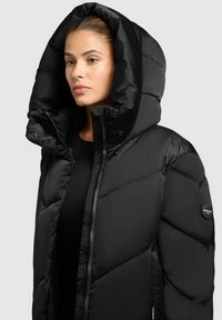 Schwarze Daunenjacke mit großer Kapuze, ausgestattet mit einem Reißverschluss und Druckknopfverschluss, gestepptem Design und runder Logo-Applikation am Ärmel.