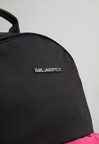 Sac à dos noir avec logo "KARL LAGERFELD" argenté et un détail en tissu rose visible en bas, sur un fond gris uni.