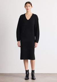 Rochie tricotată neagră cu decolteu în V, mâneci lungi și croială dreaptă. Purtați-o cu bottine negre până la gleznă. Textură netedă și design minimalist.