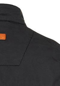 Schwarzer Kragen mit einem kontrastierenden braunen Lederetikett. Strukturiertes Gewebe mit sauberem Nähen und einer klaren Form.