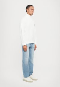 Vit zip-up sweatshirt med hög krage, i kombination med ljusblå jeans och vita sneakers, vilket visar en ren, minimalistisk design.
